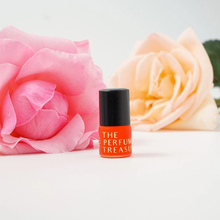 Close-up macro of floral attar perfume oil bottle by The Perfume Treasury, showing vibrant orange bottle against pink and peach roses, luxury fragrance oil in glass bottle with black cap.