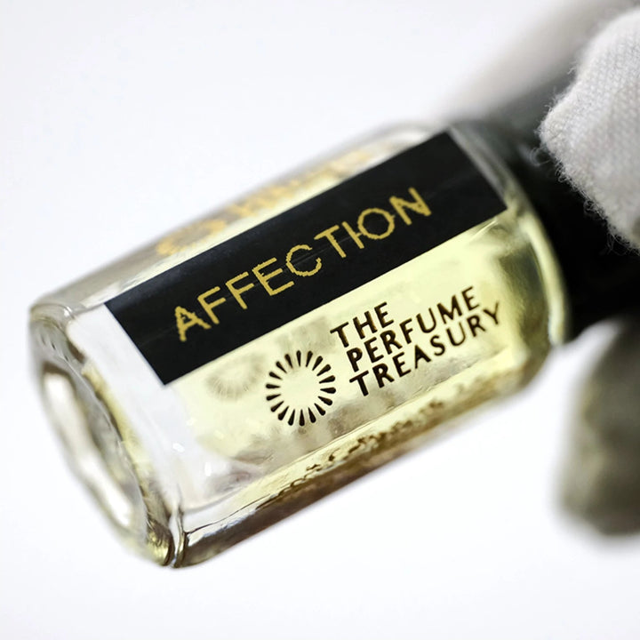 Close-up macro of Affection 3ml attar perfume oil bottle held with white glove, showing warm golden oil texture and reflective glass finish, luxury fragrance oil in clear glass bottle with black cap.