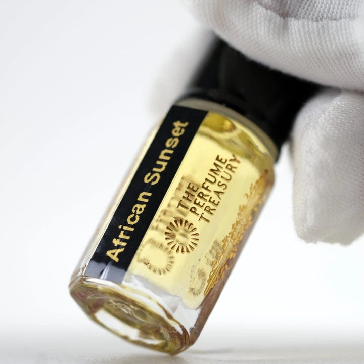 Close-up macro of African Sunset fragrance oil bottle held with white glove, showing soft golden oil texture and luxury presentation, luxury Arabian fragrance oil in clear glass bottle with black cap.