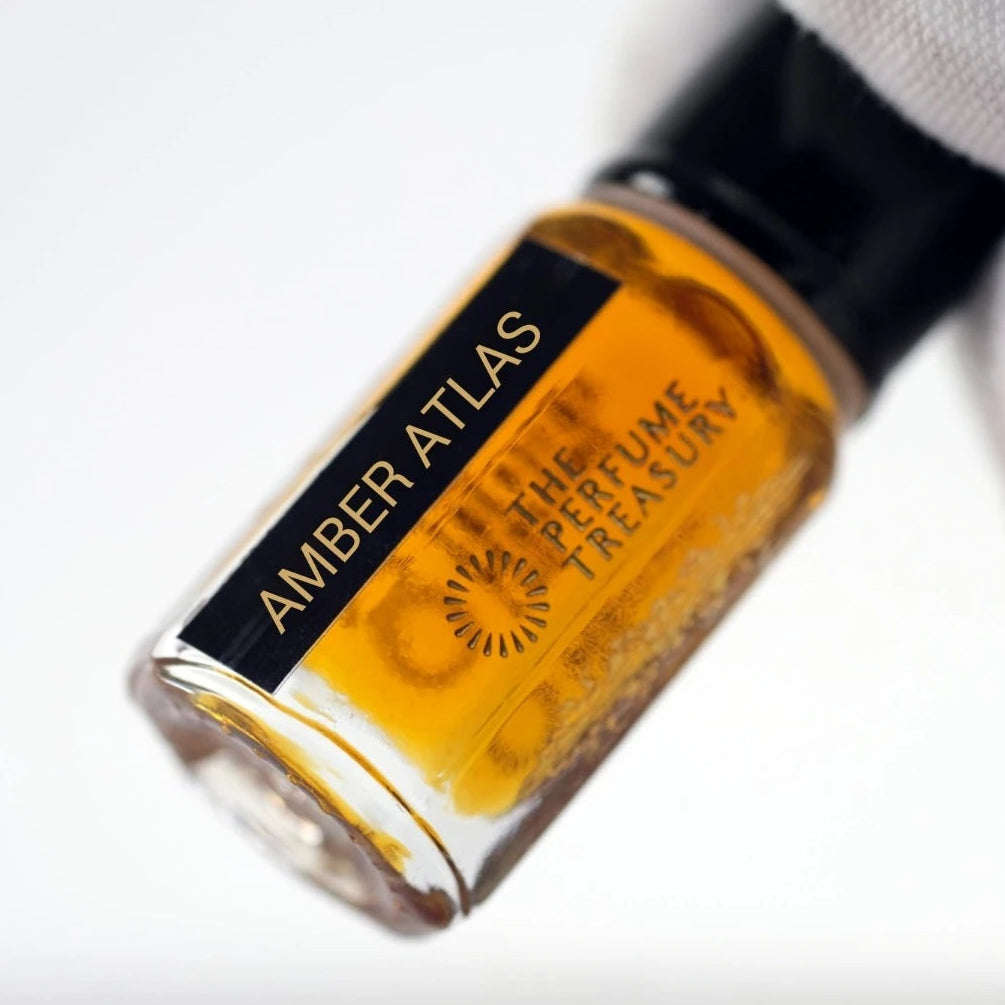 Close-up macro of Amber Atlas attar perfume oil bottle held with white glove, showing rich amber fragrance oil and luxury glass detailing, long lasting attar perfume oil in clear glass bottle with black cap.