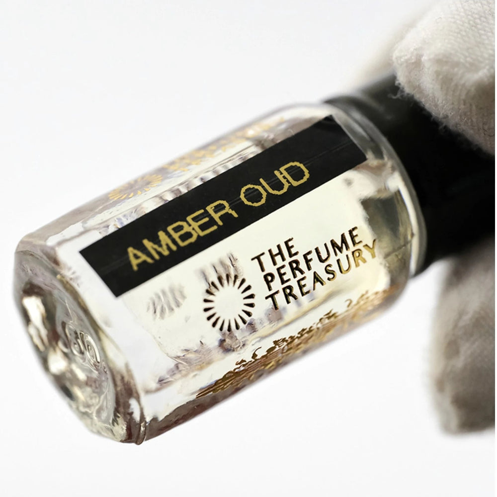 Close-up macro of Amber Oud 3ml attar perfume oil bottle held with white glove, showing glossy transparent oil texture and reflective glass finish, luxury fragrance oil in clear glass bottle with black cap.