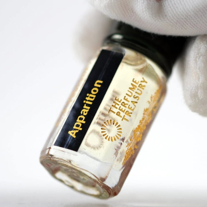 Macro detail photograph of Apparition attar fragrance oil bottle held delicately with white glove, featuring crystal-clear glass, pale golden oil and sophisticated niche perfume branding.