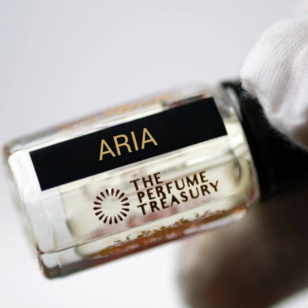 Macro close-up of Aria attar perfume bottle held in a white glove, showcasing crystal-clear fragrance oil, elegant black-and-gold label and premium glass bottle details.