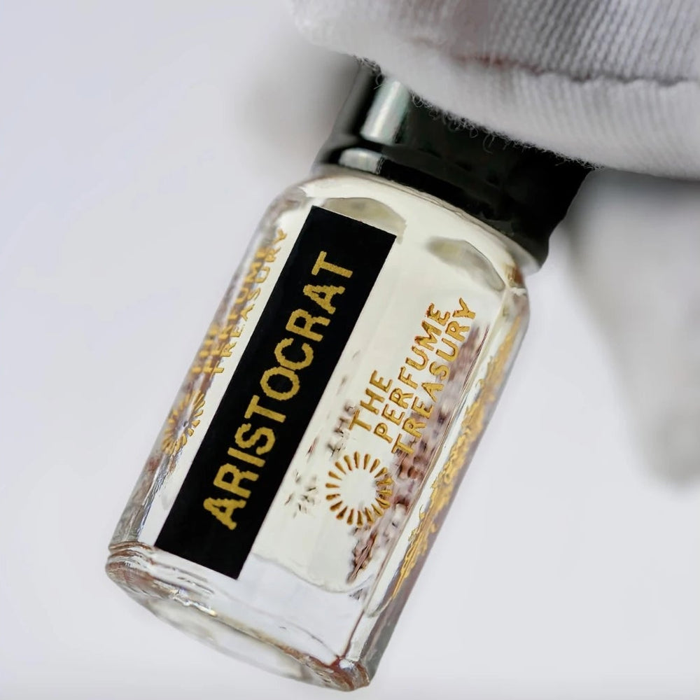 Close-up macro of Aristocrat 3ml attar perfume oil bottle held with white glove, showing clear glossy oil texture, luxury fragrance oil in clear glass bottle with black cap.