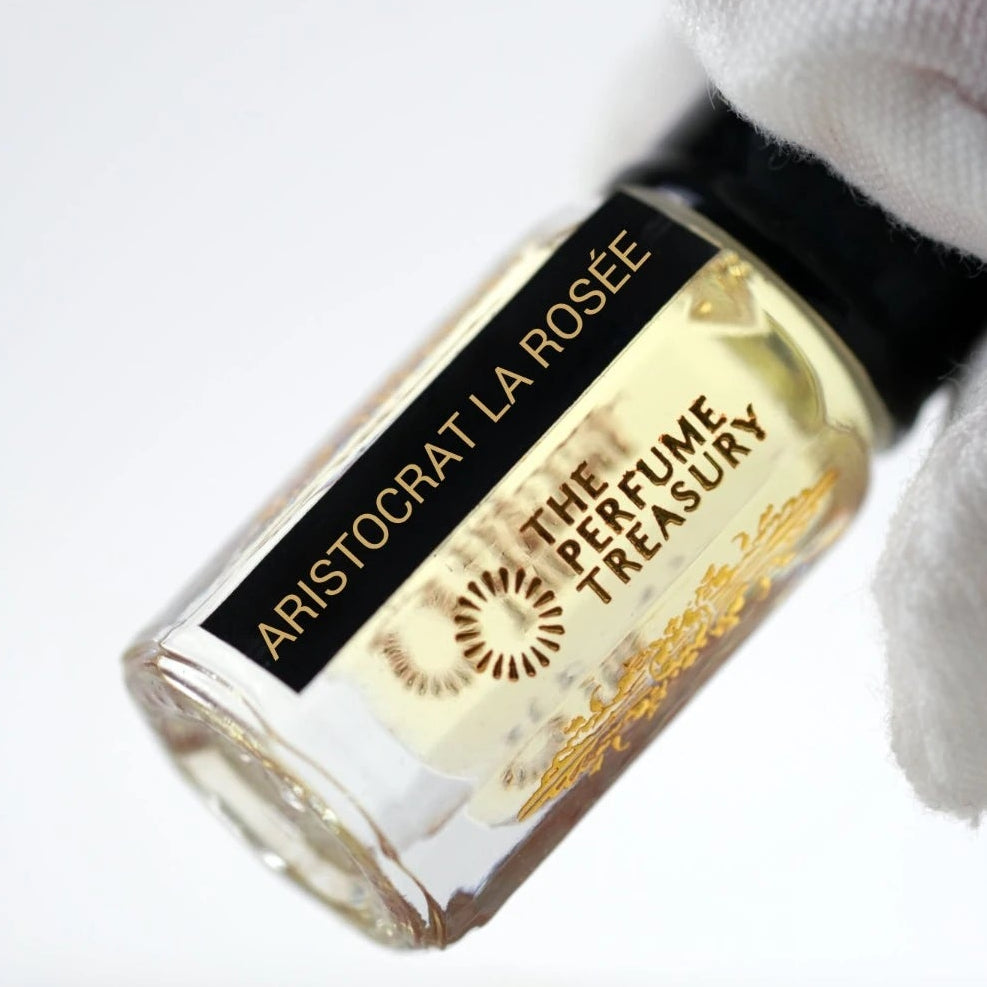Close-up luxury photograph of Aristocrat La Rosée attar perfume bottle held in a white glove, emphasizing crystal-clear fragrance oil, reflective glass and sophisticated branding details.