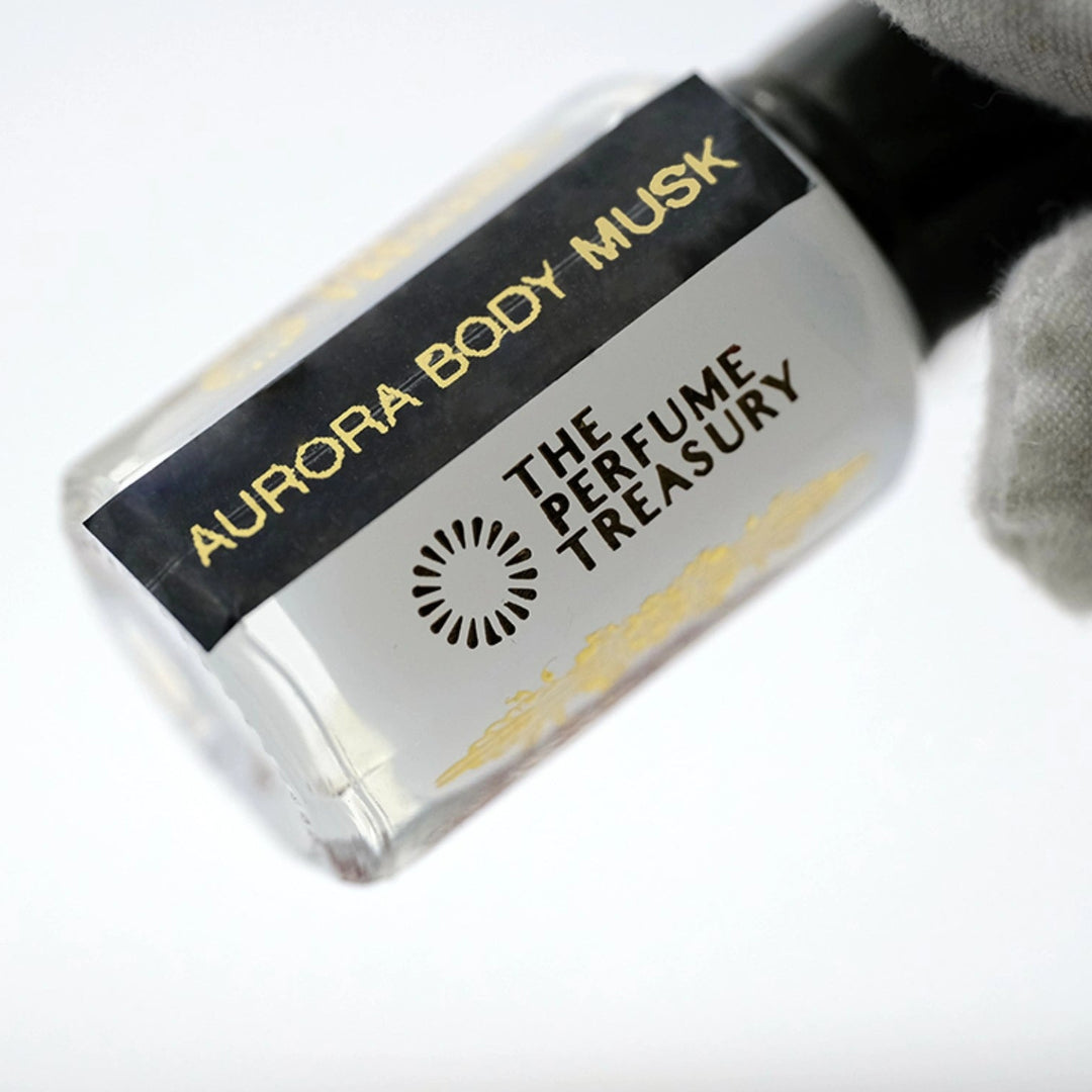Luxury close-up photograph of Aurora Body Musk attar bottle held in a white glove, highlighting the creamy white fragrance oil, sleek black label and polished glass details.