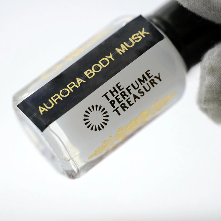 Luxury close-up photograph of Aurora Body Musk attar bottle held in a white glove, highlighting the creamy white fragrance oil, sleek black label and polished glass details.