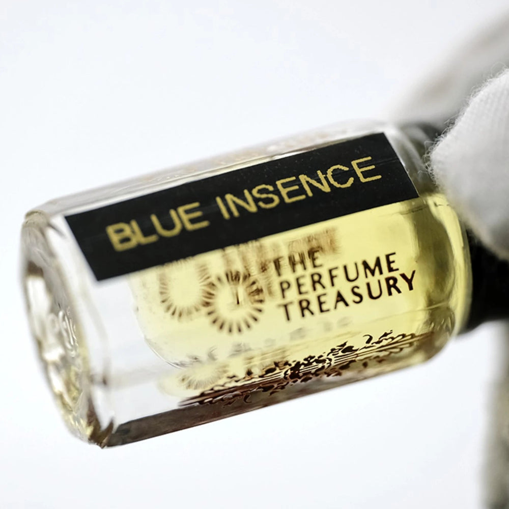Detailed close-up of Blue Insence attar bottle held in a white glove, showcasing the transparent glass, pale golden perfume oil and minimalist luxury branding.