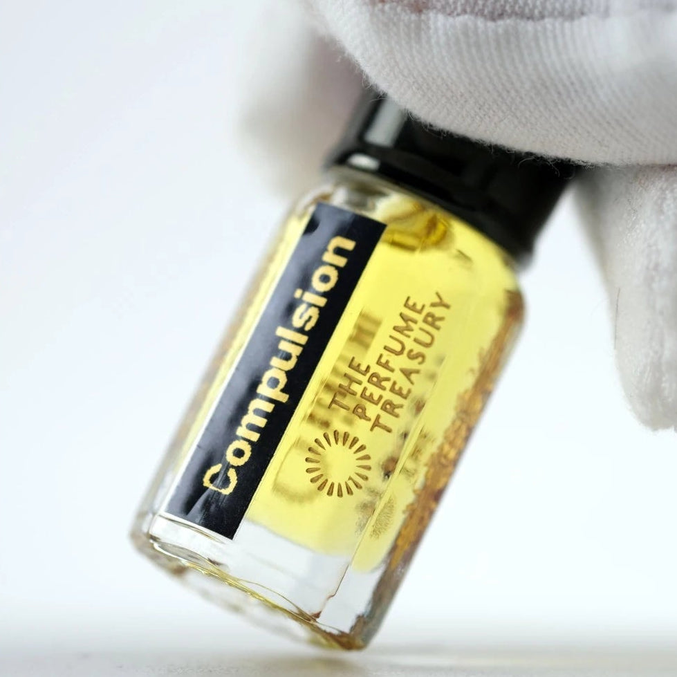 Close-up macro of Compulsion 3ml attar perfume oil bottle held with white glove, showing vibrant golden oil texture and luxury finish, luxury fragrance oil in clear glass bottle with black cap.