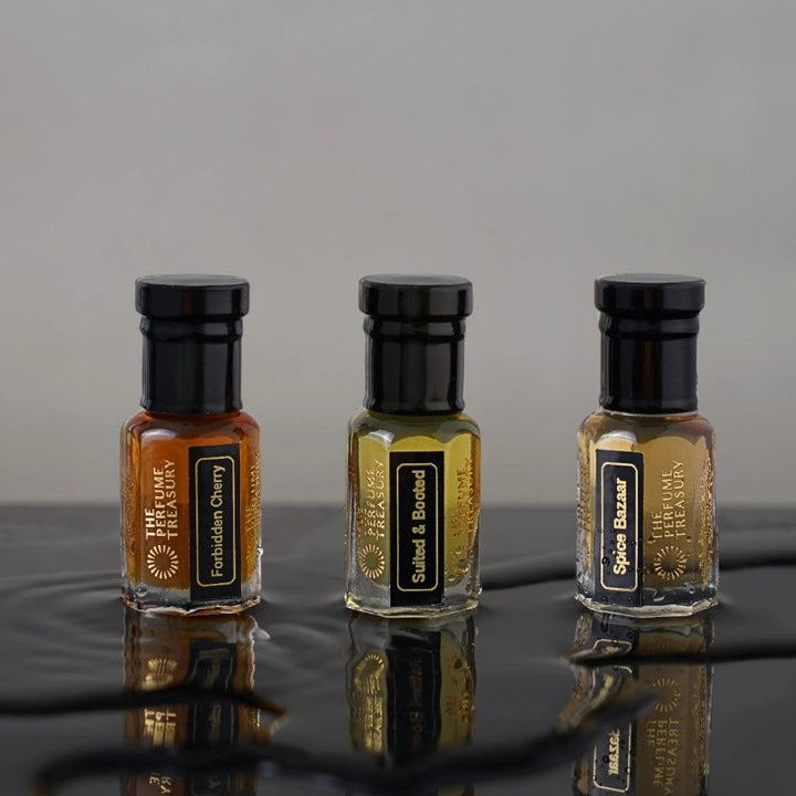 Small clear glass perfume oil bottles with black caps, Forbidden Cherry, Sultry &amp; Booted and Spice Bazaar 3ml attar perfume oils by The Perfume Treasury, rich amber and golden fragrance oils on reflective black surface.