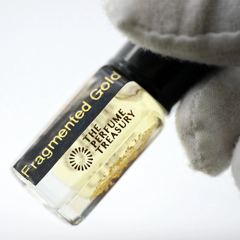 Close-up of Fragmented Gold attar perfume oil by The Perfume Treasury, showcasing the pale golden fragrance oil, black label and decorative gold glass bottle detailing.