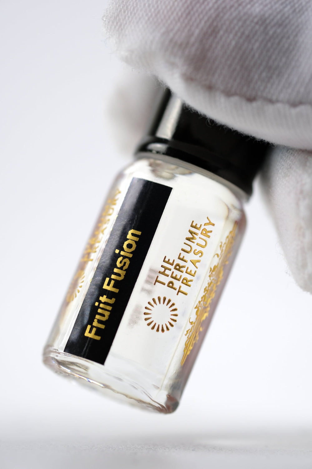 Close-up macro of Fruit Fusion 3ml attar perfume oil bottle by The Perfume Treasury, showing glossy smooth fragrance oil texture and gold detailing, luxury oil in clear bottle with black cap.