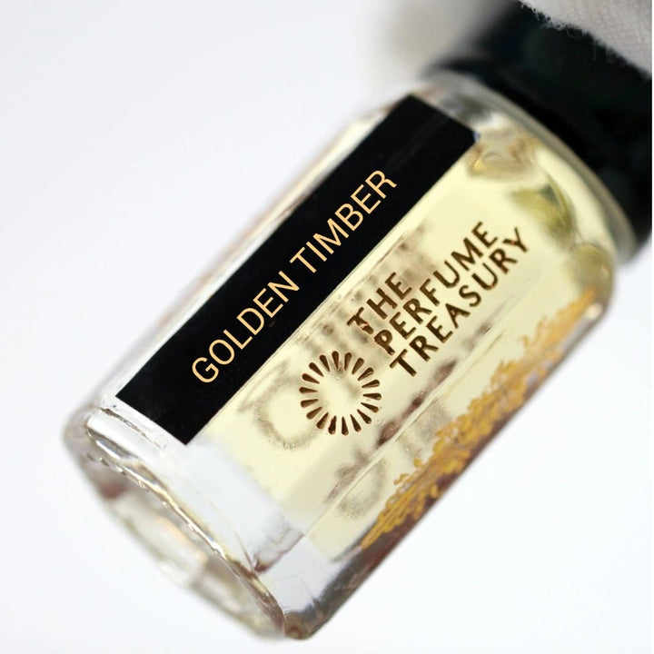 Close-up of a small Golden Timber Attar Perfume Oil bottle by The Perfume Treasury, 3ml roll-on perfume oil with black cap and black and gold label, photographed against a soft white background.
