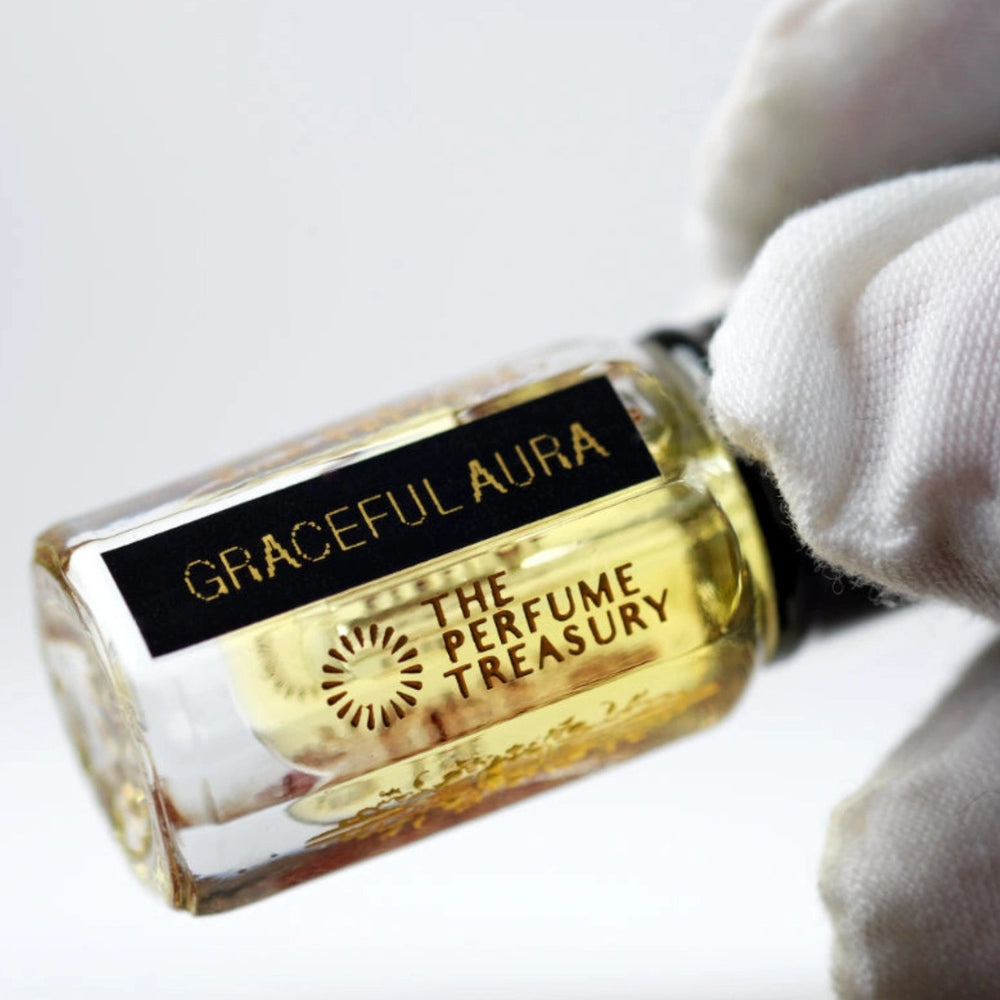 A small clear Roll-On perfume bottle filled with golden liquid, held by a white gloved hand. The bottle has a black label with gold text reading 'GRACEFUL AURA' and 'THE PERFUME TREASURY'.