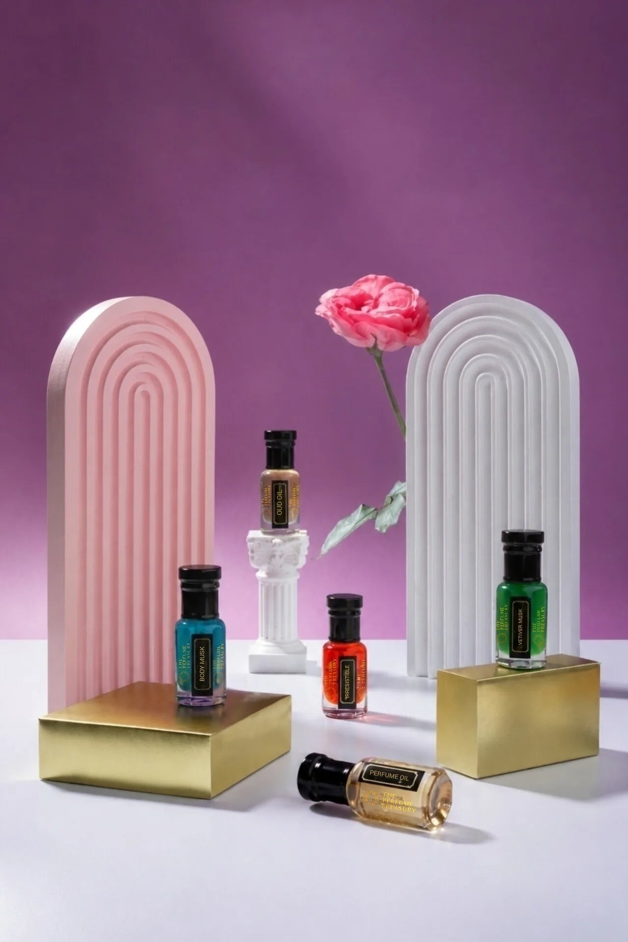 The Perfume Treasury perfume oil bottles styled on gold pedestals with pink arches and a rose on a purple background