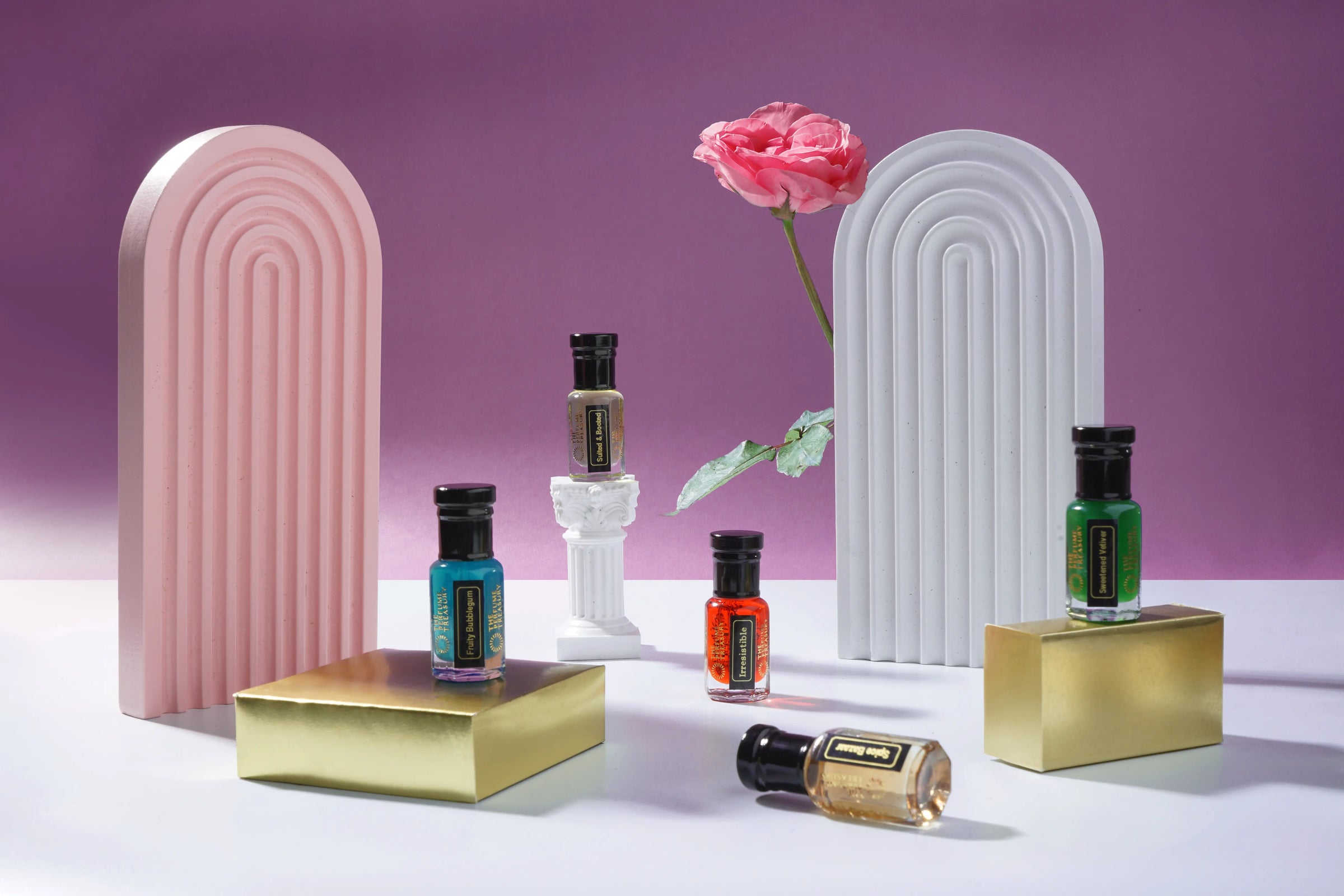 The Perfume Treasury perfume oil bottles styled on gold pedestals with pink arches and a fresh rose