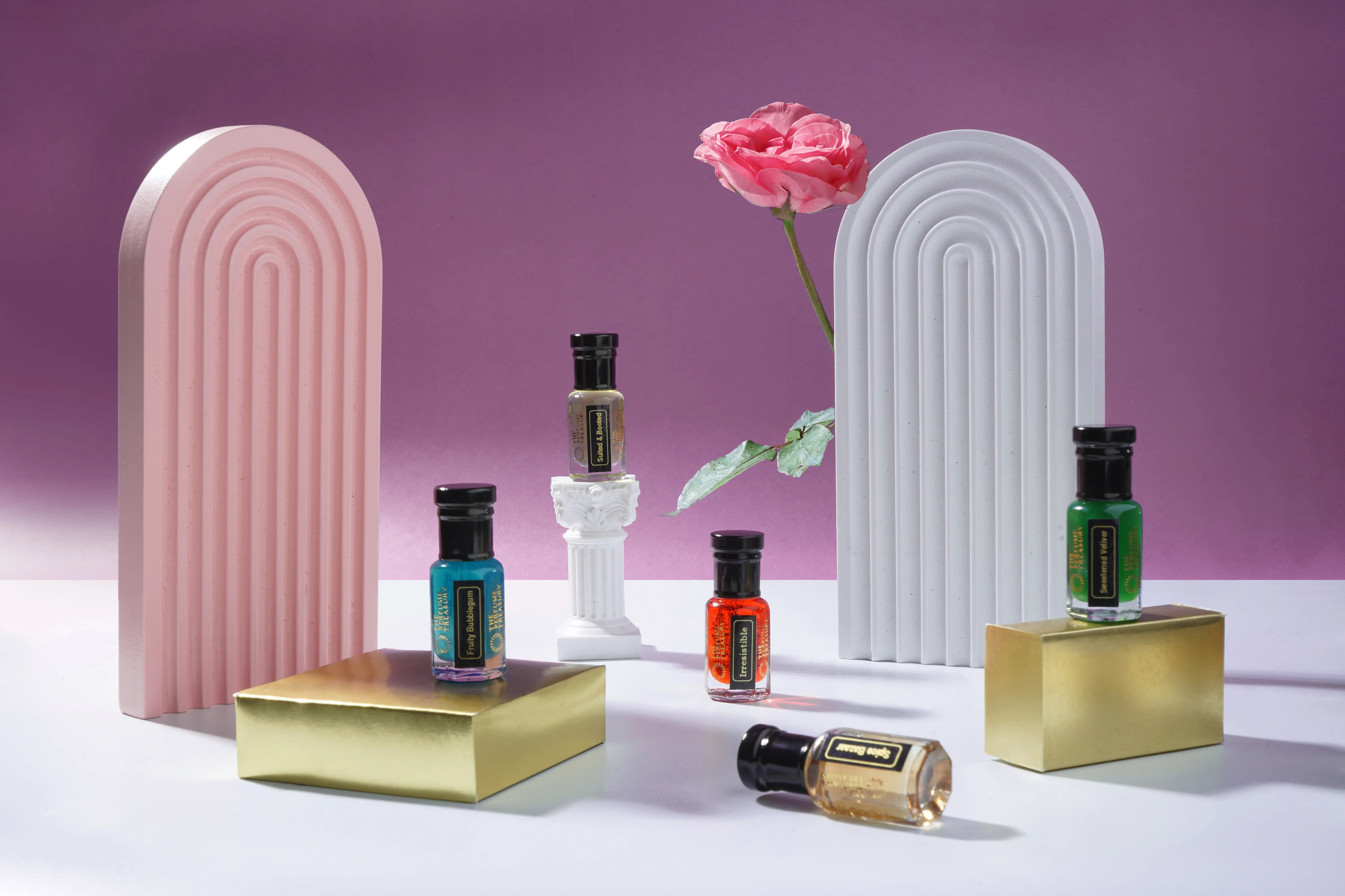 The Perfume Treasury perfume oil bottles styled on gold pedestals with pink arches and a fresh rose