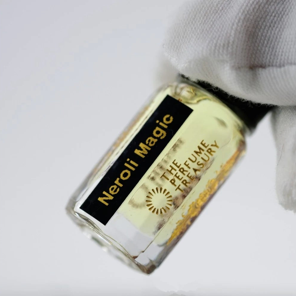 Close-up of Neroli Magic perfume oil by The Perfume Treasury held in a white glove, showing the black and gold label and pale golden fragrance oil.
