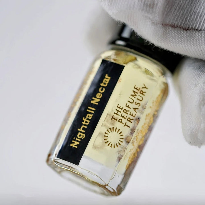 Close-up of Nightfall Nectar perfume oil by The Perfume Treasury held in a white glove, showing the black and gold label and golden fragrance oil.