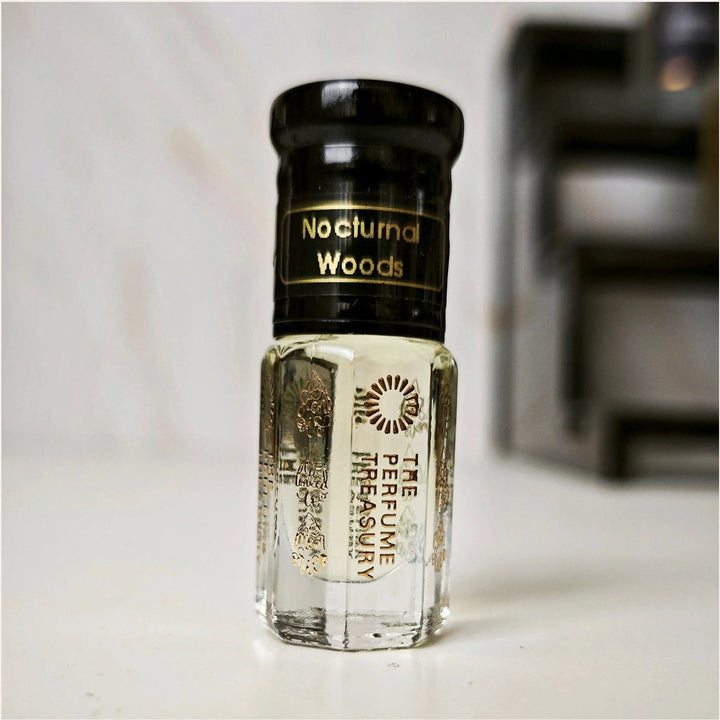 Small clear glass perfume oil bottle with black cap, Nocturnal Woods 3ml attar perfume oil by The Perfume Treasury, clear to pale golden fragrance oil on soft neutral background.
