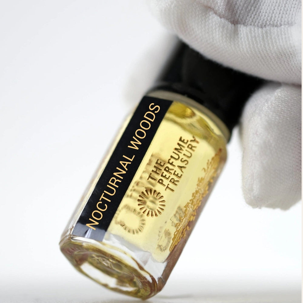 Close-up of small clear glass perfume oil bottle with black cap, Nocturnal Woods 3ml attar perfume oil by The Perfume Treasury, golden fragrance oil held in white gloved hand on white background.