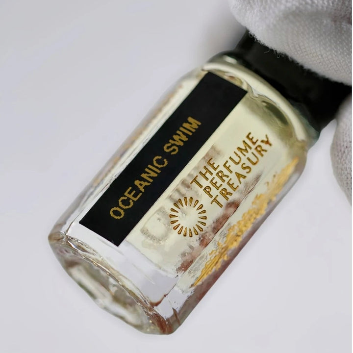 Close-up of Oceanic Swim perfume oil by The Perfume Treasury held in a white glove, showing the black and gold label and pale golden fragrance oil.