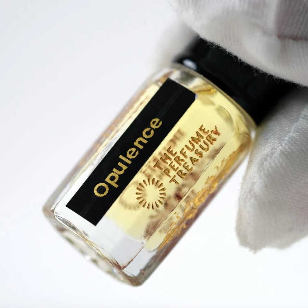 Close-up of Opulence perfume oil by The Perfume Treasury held in a white glove, showing the black and gold label and pale golden fragrance oil.