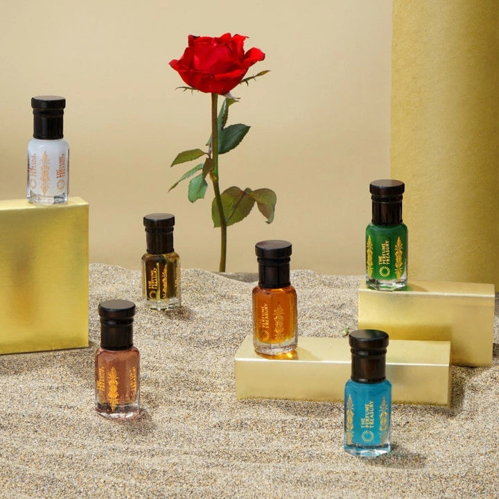 Luxury display of multiple perfume oil bottles by The Perfume Treasury with coloured fragrance oils, arranged on gold blocks with red rose on textured sand background. 