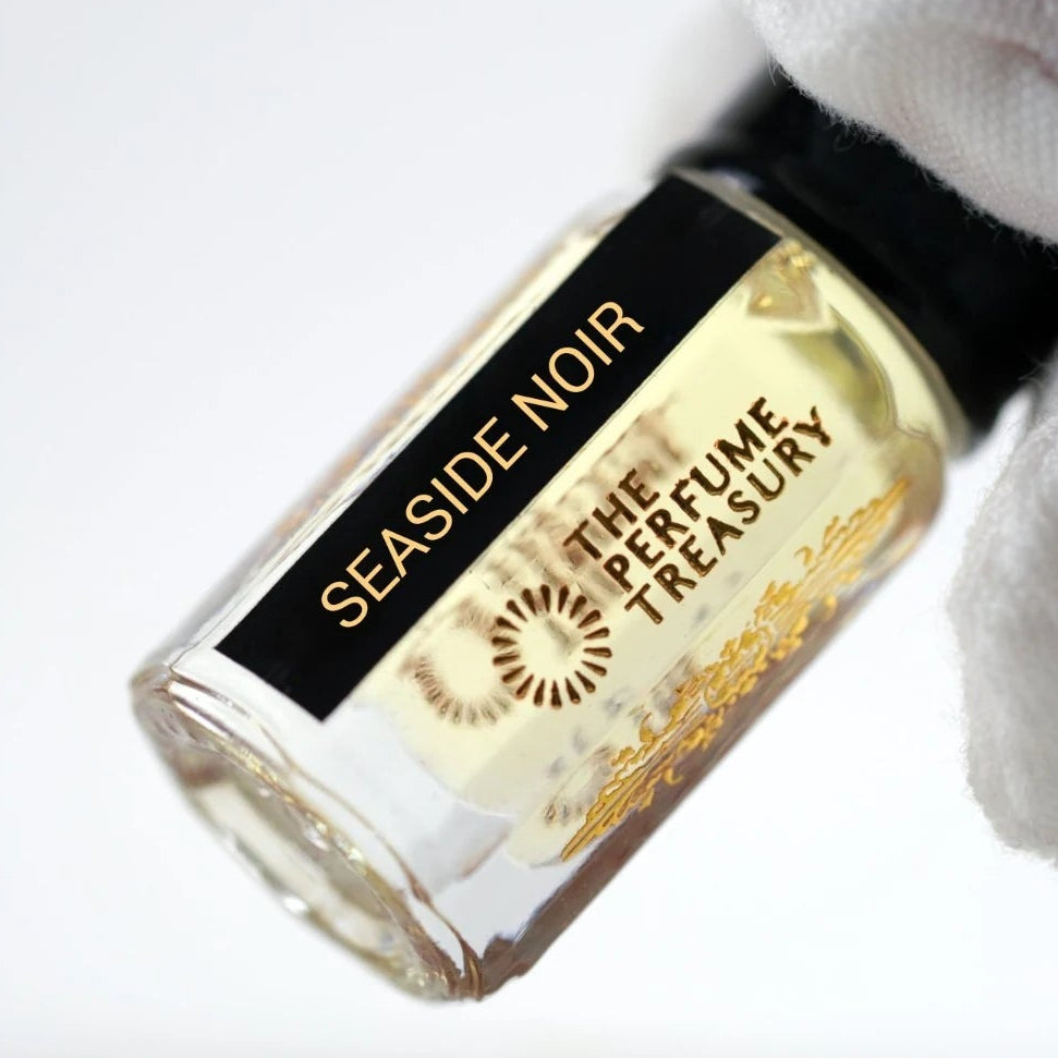 Seaside Noir perfume oil by The Perfume Treasury held in a white-gloved hand, showcasing the clear glass bottle and pale golden fragrance oil.