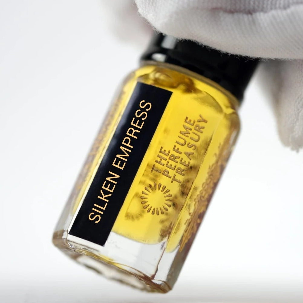 Silken Empress perfume oil inspired by Creed Queen of Silk in a clear glass bottle filled with golden fragrance oil, with a black lid and gold lettering, held by a white-gloved hand