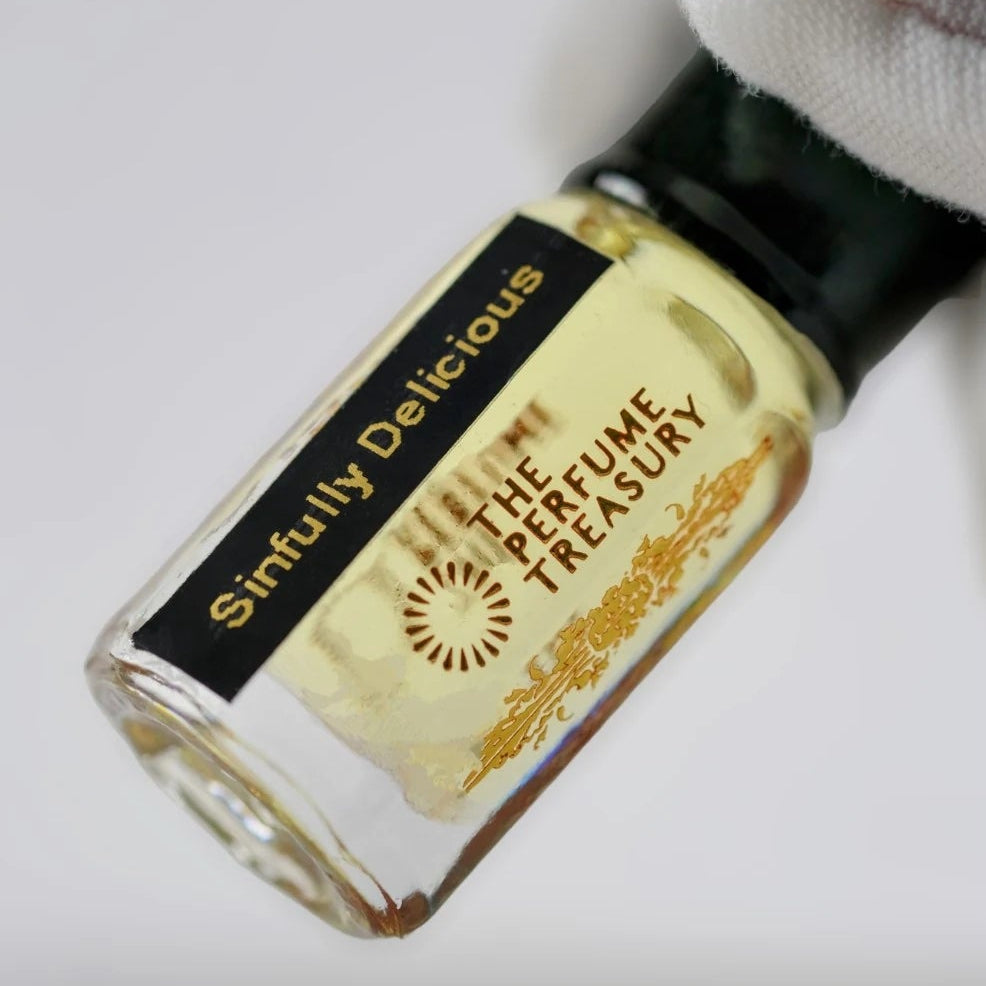 Sinfully Delicious perfume oil inspired by Montale Chocolate Greedy in a clear glass bottle with pale golden fragrance oil, black lid and black gold label, held by a white-gloved hand