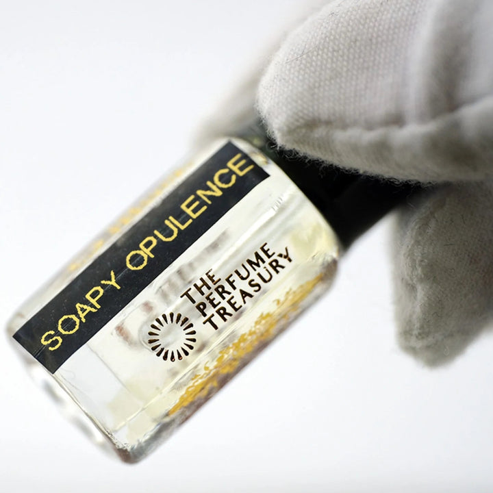 Soapy Opulence perfume oil by The Perfume Treasury in a clear glass bottle with pale fragrance oil, glossy black lid and black gold label, held at an angle by a white-gloved hand