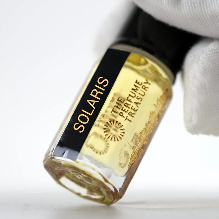 Solaris perfume oil by The Perfume Treasury in a clear glass bottle with golden fragrance oil, glossy black lid and black gold label, held at an angle by a white-gloved hand against a clean white background