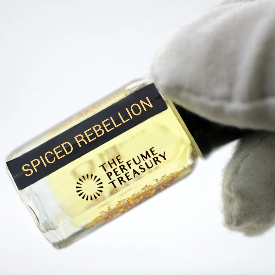 Spiced Rebellion perfume oil by The Perfume Treasury in a clear glass bottle with golden fragrance oil, black lid and black gold label, held at an angle by a white-gloved hand