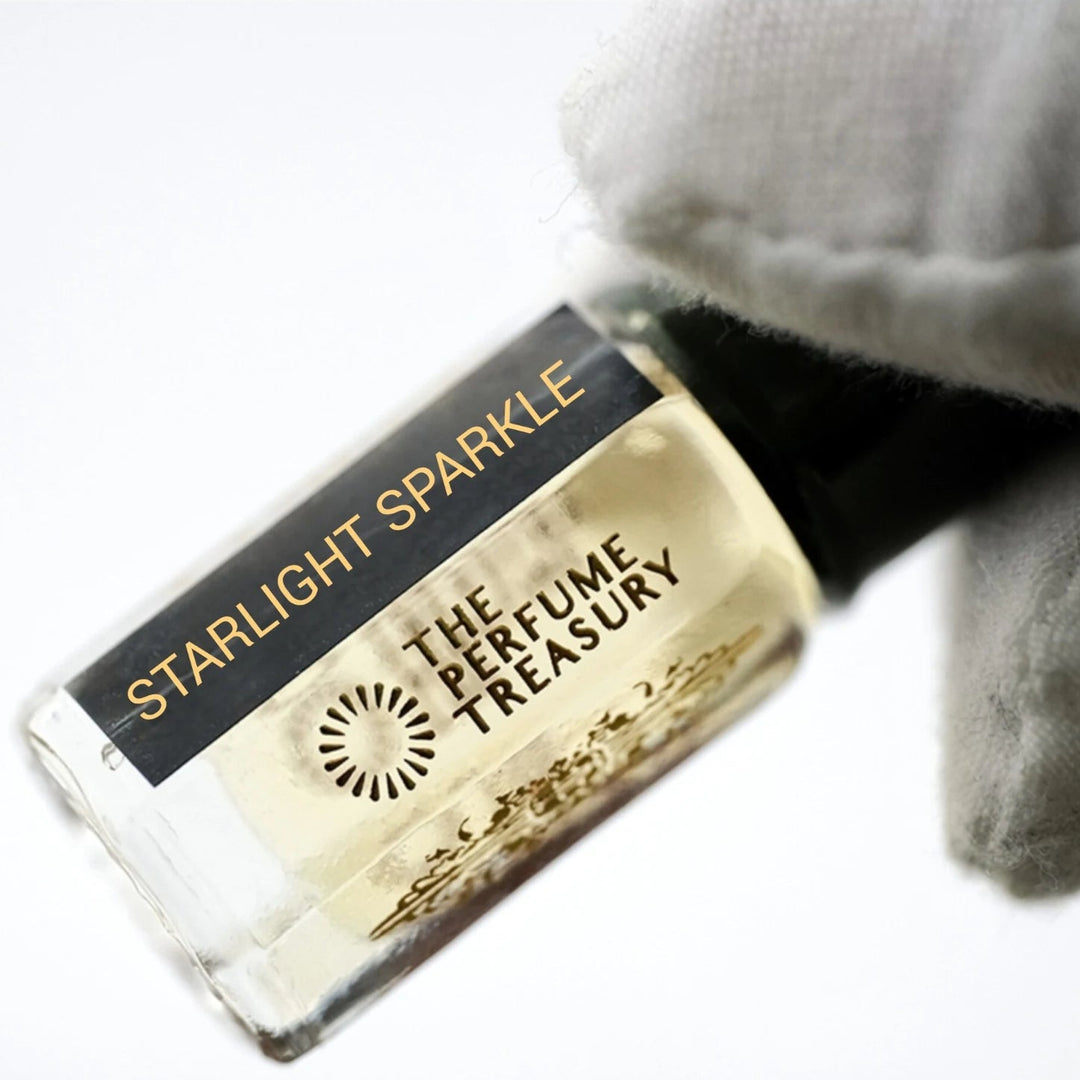 Starlight Sparkle perfume oil by The Perfume Treasury in a clear glass bottle with pale golden fragrance oil, black lid and black gold label, held by a white-gloved hand