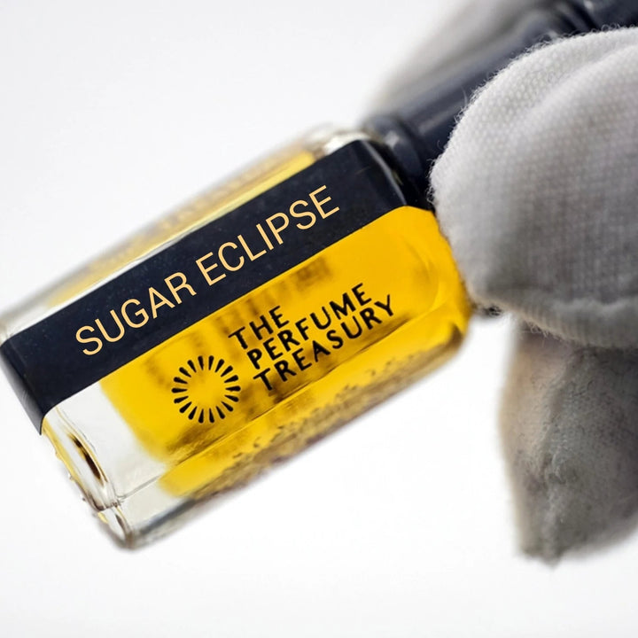 Sugar Eclipse perfume oil by The Perfume Treasury with rich golden fragrance oil, black lid and black gold label, held at an angle by a white-gloved hand