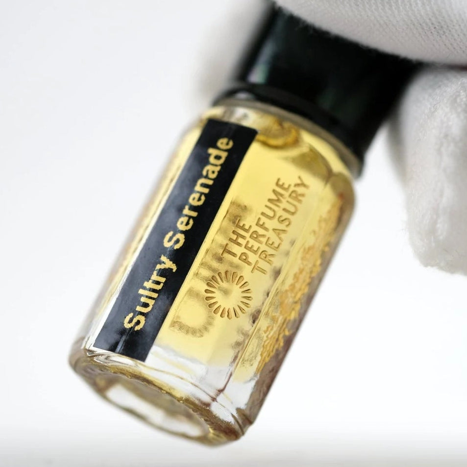 Sultry Serenade perfume oil by The Perfume Treasury in a clear glass bottle with golden fragrance oil and black gold label, held at an angle by a white-gloved hand