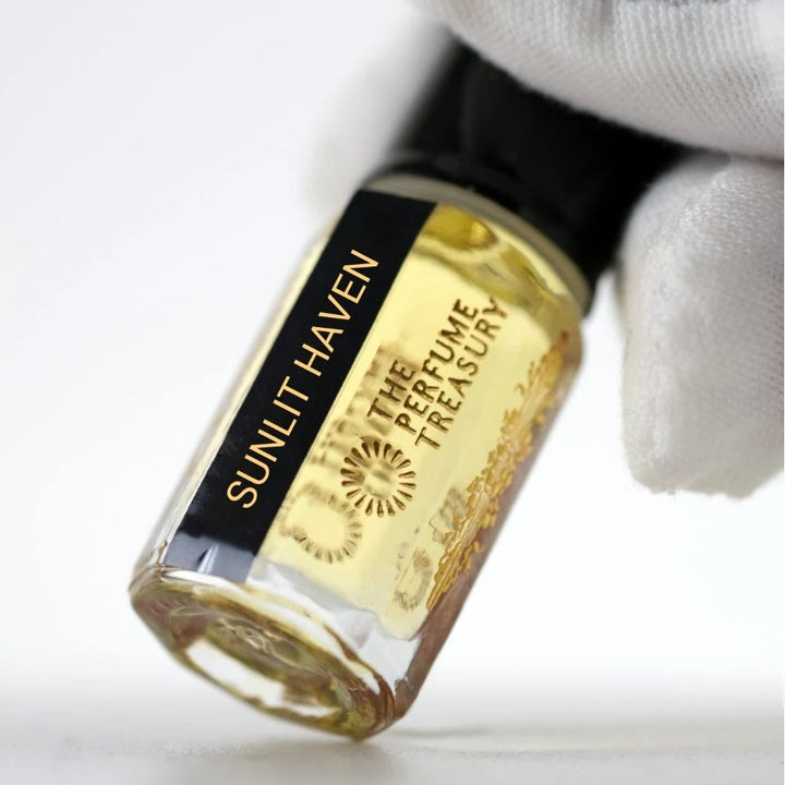 Sunlit Haven perfume oil by The Perfume Treasury in a clear glass bottle with pale golden fragrance oil and black gold label, held at an angle by a white-gloved hand