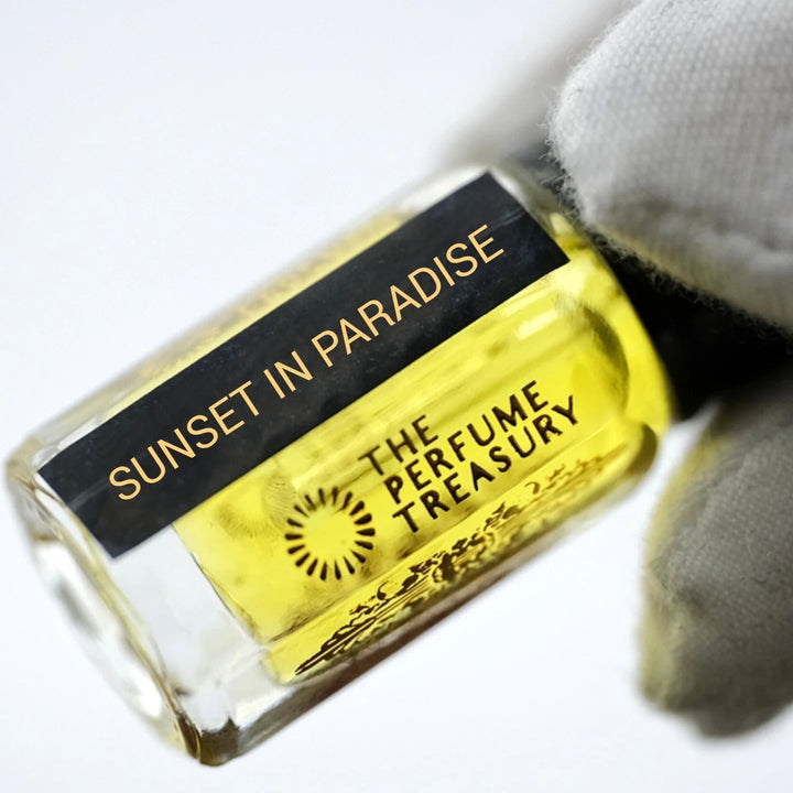 Sunset in Paradise perfume oil by The Perfume Treasury in a clear glass bottle with golden fragrance oil and black gold label, held at an angle by a white-gloved hand